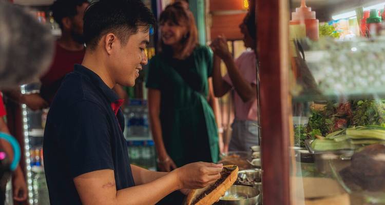 Un vendedor de comida callejera prepara un sándwich mientras los turistas del grupo observan y charlan detrás del puesto
