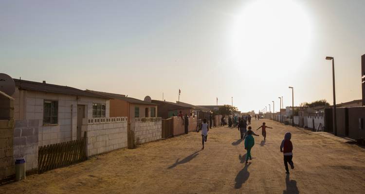 Des enfants courent le long d'une rue poussiéreuse du township vers le soleil couchant bas, avec de simples maisons qui bordent la route.