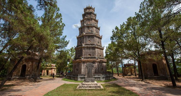 La pagode historique Thien Mu s'élève au milieu des pins à Hué avec des visiteurs marchant sur le chemin de briques ci-dessous.