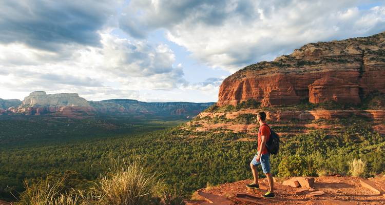 Jeune randonneur se tenant debout sur une falaise de roche rouge à Sedona surplombant une vaste vallée verte et des mesas de grès.