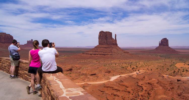 Toeristen die foto's maken van imposante zandstenen tafelbergen bij het uitkijkpunt van Monument Valley onder een blauwe lucht.