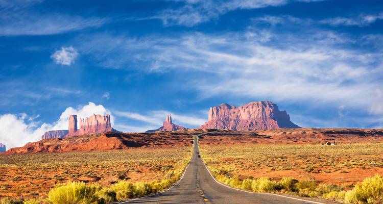 Carretera infinita y recta que lleva hacia las enormes mesetas rojas de Monument Valley bajo un cielo espectacular.