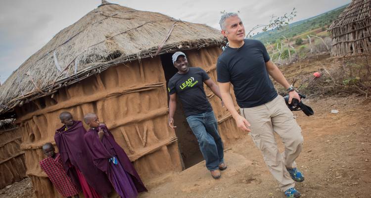 A traveller exits a traditional Maasai hut while locals in purple robes stand at the doorway.