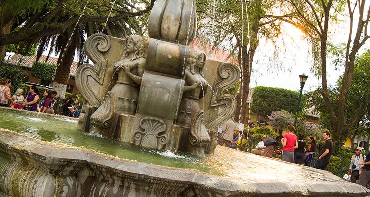 Una ornada fuente de piedra con figuras de dos sirenas salpica en una animada plaza colonial.