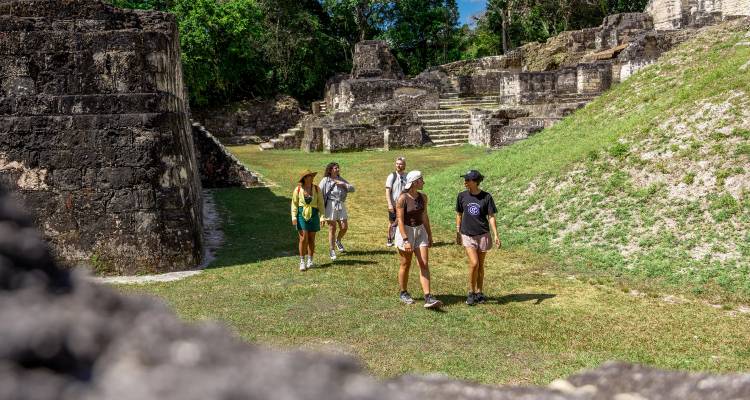 Les voyageurs marchent parmi les anciennes structures de pierre maya et les terrasses herbeuses sous un soleil éclatant.