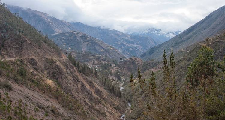 Vallée andine profonde avec une route de terre sinueuse, une rivière et des pics couverts de nuages s'étendant à l'horizon.