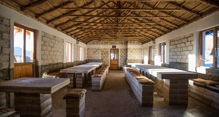 Comedor construido en piedra con mesas de bloques de sal largas y asientos de banco iluminados por ventanas laterales.