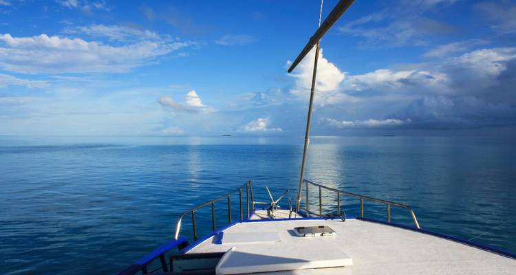 Proue d'un bateau fendant une mer calme et ouverte sous un ciel bleu brillant avec des nuages épars.