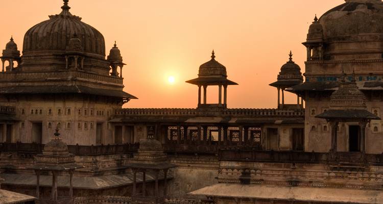 Los dómes y chattris silueteados de un palacio histórico brillan bajo una puesta de sol anaranjada.