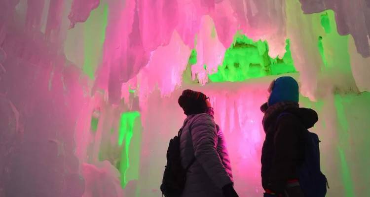 Groepjes bezoekers kijken omhoog naar roze en groen verlichte ijsformaties in een gloeiende ijsgrot.