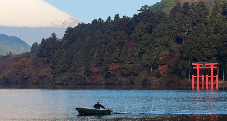 Une petite embarcation glisse sur le lac Ashi avec le torii rouge parmi la rive boisée et le Mont Fuji enneigé au-delà.