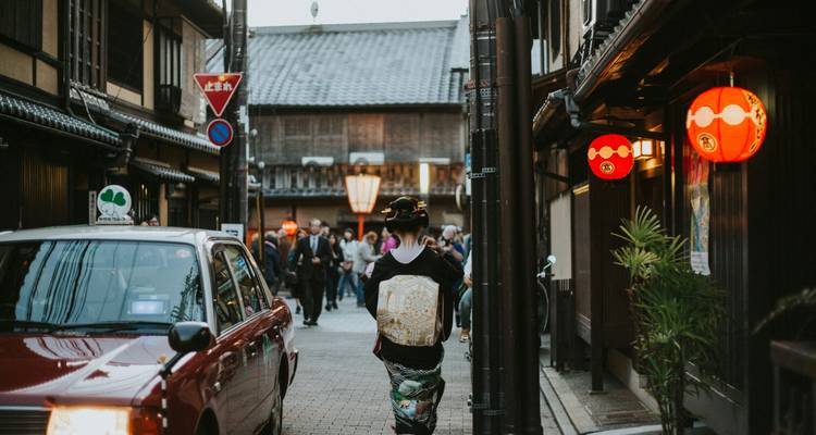 Traditional lantern-lit alley in Kyoto with a kimono-clad woman walking past a taxi while crowds stroll between wooden machiya houses
