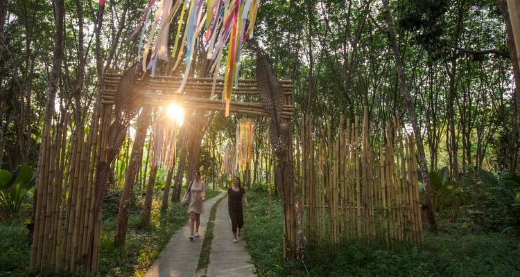 Deux femmes se promènent sur un étroit sentier sous une arche en bambou décorée de rubans colorés au coucher du soleil.