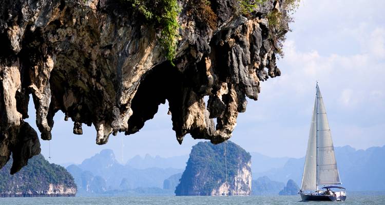 Un yacht navigue devant d'imposantes formations calcaires de type karst sous une formation rocheuse surplombante sur des eaux tranquilles.