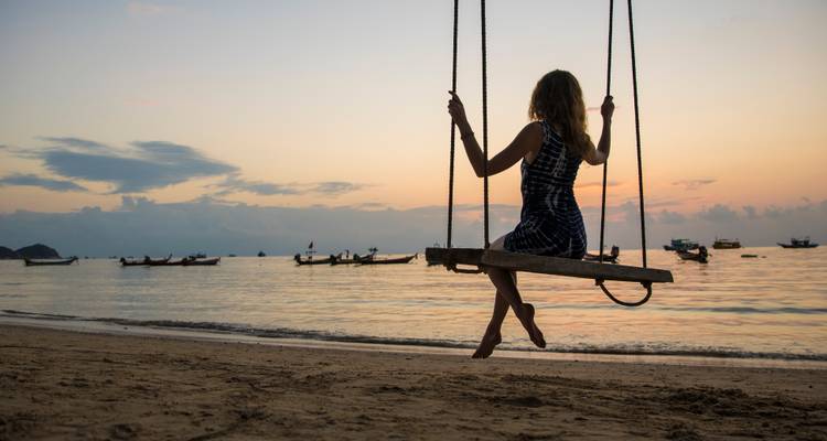 Mujer en silueta sentada en un columpio de madera sobre aguas tranquilas del mar en una puesta de sol pastel.