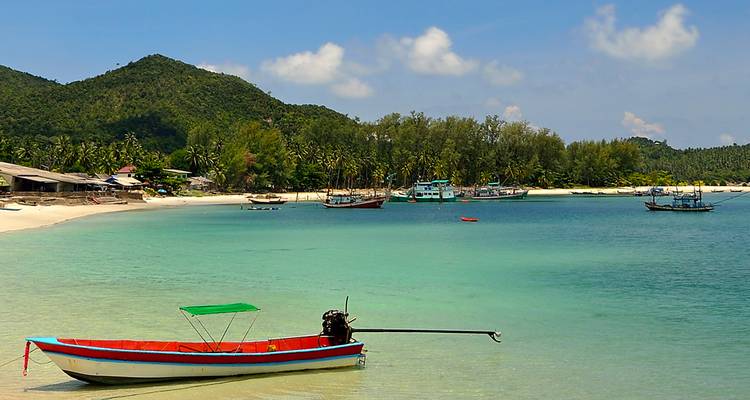 Bahía tropical de agua turquesa con un colorido bote de cola larga amarrado frente a una playa de arena blanca y colinas verdes al fondo
