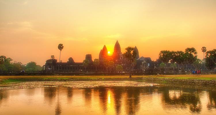 Angkor Wat en silhouette lors d'un lever de soleil orange chaleureux, parfaitement reflété dans un étang immobile bordé de palmiers.