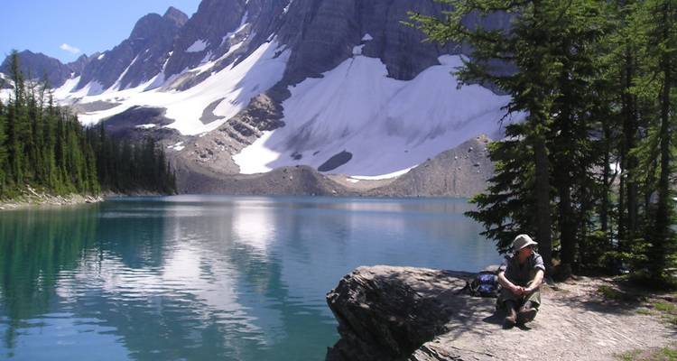 Persona sentada junto a un lago sereno con montañas cubiertas de nieve.