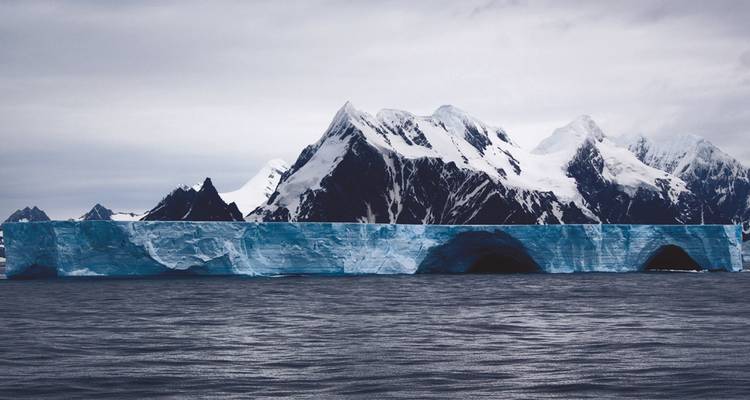 Ein massiver, flachgipfeliger Eisberg erstreckt sich über grauem antarktischem Wasser mit gezackten Gipfeln dahinter