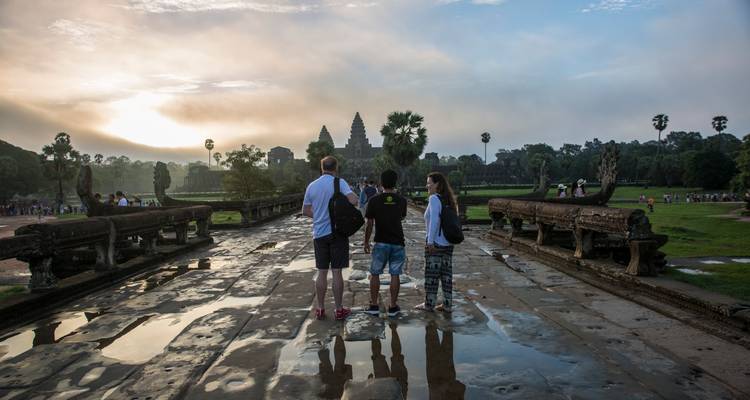 Drei Reisende stehen auf einem reflektierenden Steinweg und bewundern Angkor Wat bei Sonnenaufgang, während goldenes Licht durch die Wolken bricht.