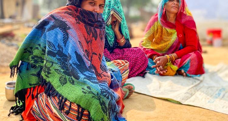 Women wrapped in brightly coloured shawls sit on the ground in a rural Indian setting.