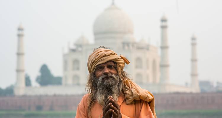 A bearded sadhu poses with hands together, with the Taj Mahal softly visible in the background.