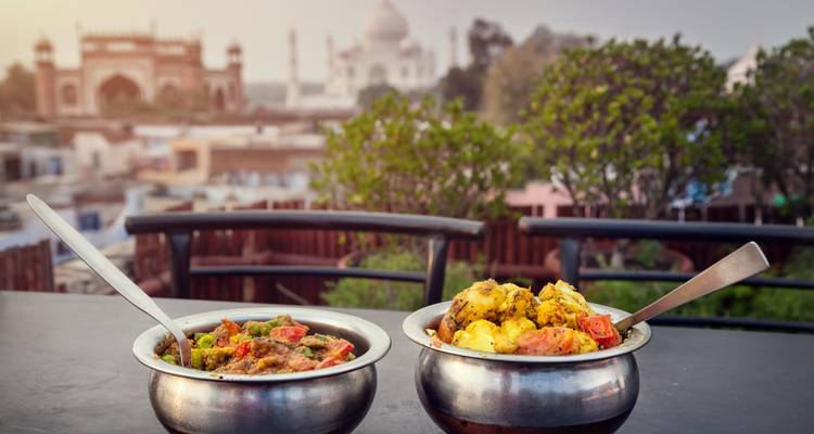 Two metal bowls of colourful Indian curry on a rooftop with the Taj Mahal blurred in the distance.