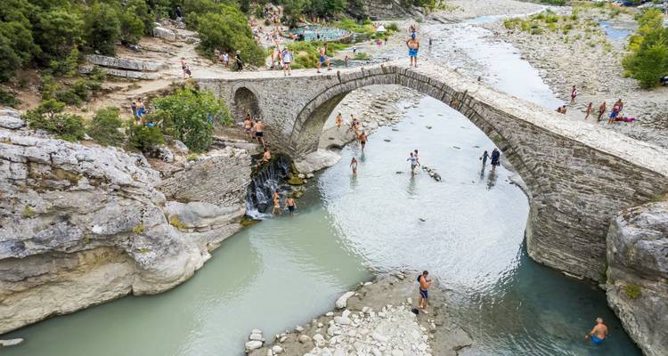 Historische stenen boogbrug over türkooizen rivier met veel zwemmers die genieten van natuurlijke zwembaden eronder.
