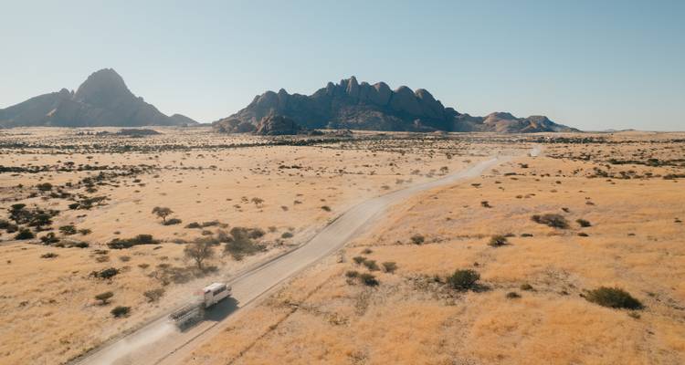 Vue aérienne d'un camion de safari solitaire soulevant la poussière sur une route de terre à travers les plaines désertiques dorées en direction des pics dentelés de Spitzkoppe.