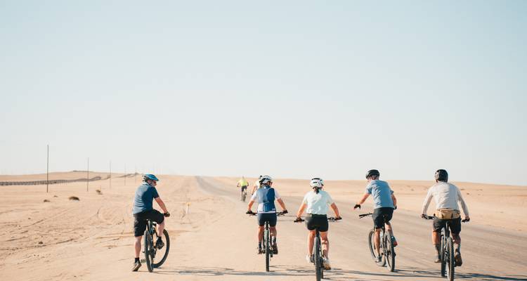 Les cyclistes roulent en file le long d'une route désertique rectiligne sous un ciel bleu pâle.