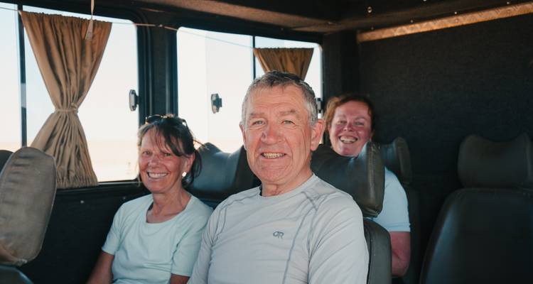Trois voyageurs d'un certain âge sourient en étant assis à l'intérieur d'un camion de voyage avec un paysage désertique visible à travers les fenêtres.