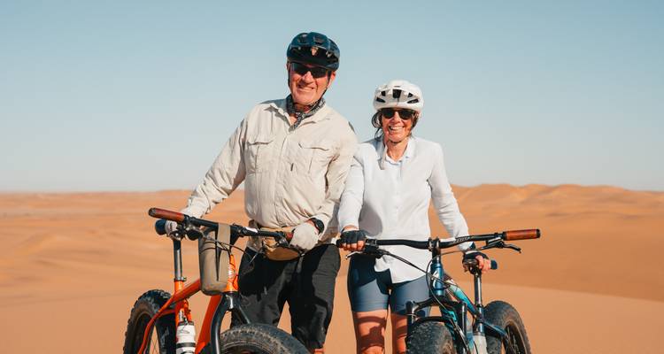 Un couple souriant se tient debout avec des vélos à gros pneus au sommet des dunes dorées du désert sous un ciel dégagé.