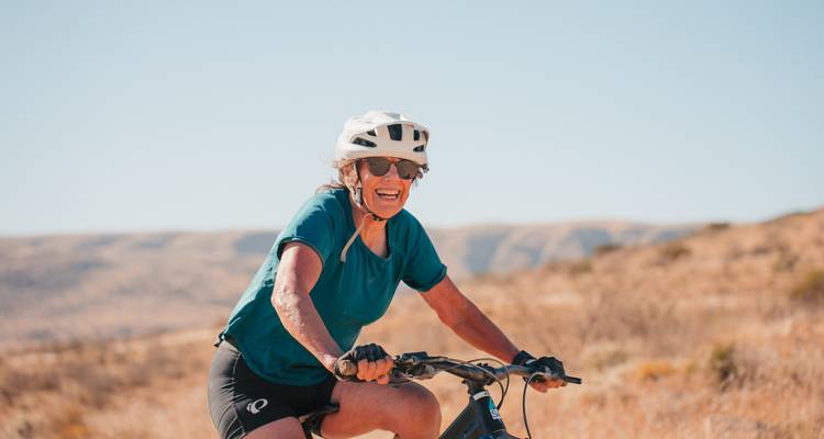 Une femme portant un casque rit en faisant du vélo à travers des prairies dorées et arides par une belle journée ensoleillée.