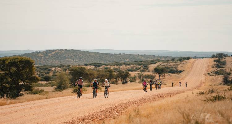 Une ligne de cyclistes s'étend le long d'une route en gravier rougeâtre traversant un paysage de brousse ouvert.