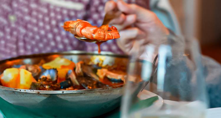 Primer plano de un guiso de mariscos con una mano levantando un camarón del plato, con una copa desenfocada en primer término.