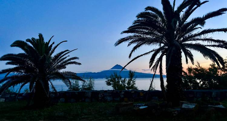 Los árboles de palma recortados enmarcan un océano tranquilo con un volcán de forma cónica a lo lejos durante el atardecer.