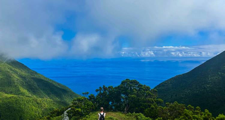 Un senderista solitario se encuentra de pie en una cresta verde que domina un mar azul profundo entre las laderas de islas cubiertas de niebla.