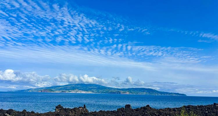 Orilla rocosa de lava enfrenta una isla verde distante bajo nubes rayadas y cielo azul brillante.