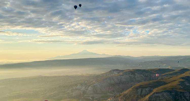 Suave luz matinal sobre valles brumosos, volcán a lo lejos y globos aerostáticos dispersos por el cielo.
