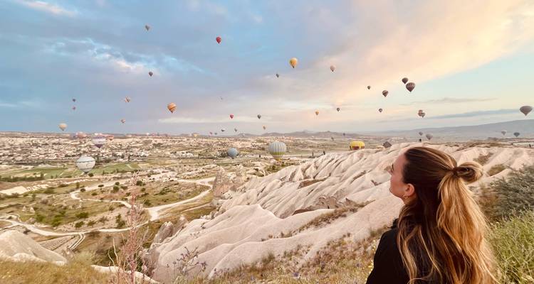 Una mujer admira docenas de globos elevándose sobre las crestas rocosas de tonos pastel de Capadocia al amanecer.