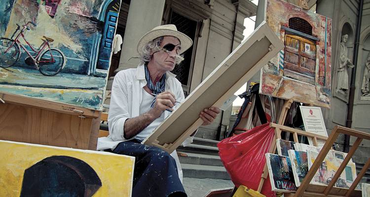 Artista callejero sentado en un caballete dibujando en una plaza italiana rodeado de cuadros.