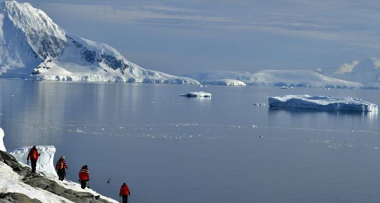 Travellers in red parkas trek along a rocky Antarctic shoreline with calm reflective water and floating icebergs beneath a snowy mountain backdrop.