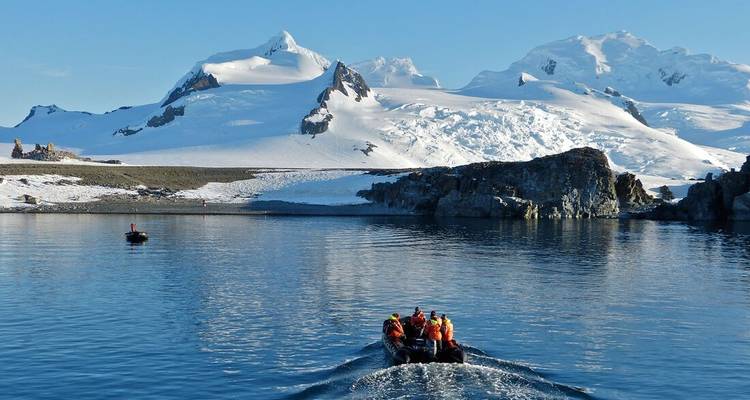 Zodiac boat full of tourists speeds toward a snow-covered Antarctic island with jagged peaks reflecting on glassy water.