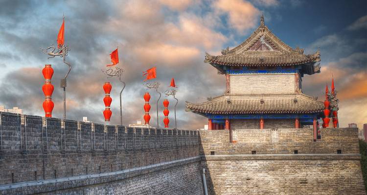 De stadsmuur van Xi'an met bastions versierd met rode lantaarns en banieren tegen een dramatische zonsonderganglucht.