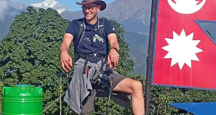 Un trekker sonriente frente a una bandera de Nepal está de pie en un mirador con colinas verdes y picos.