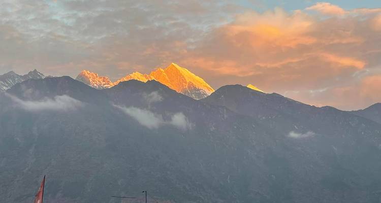 Golden sunrise light touches a Himalayan peak while low clouds drift below and sky glows orange.