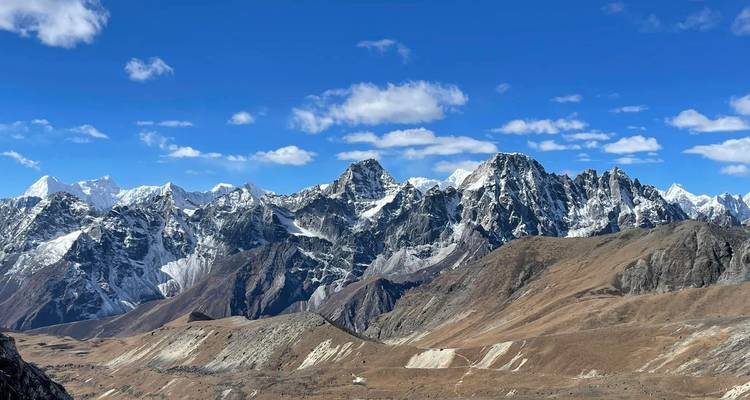 Wide panorama of jagged grey Himalayan ridges and snow patches under a deep blue sky with scattered clouds.