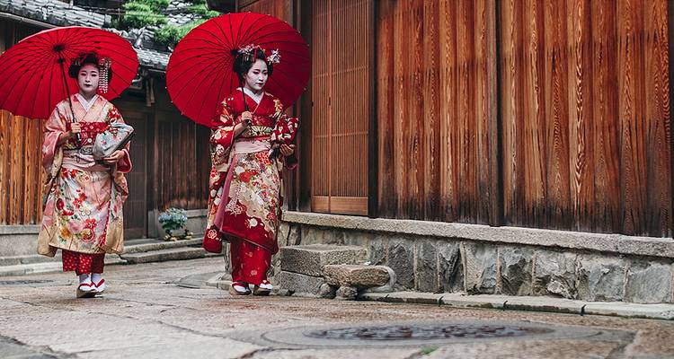Zwei Personen in traditioneller japanischer Kleidung, die rote Regenschirme halten, gehen eine Straße mit Holzhäusern entlang.
