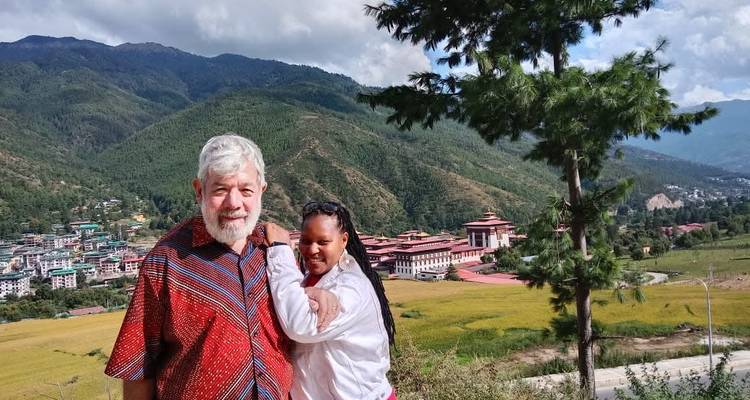 An older man and younger woman smile on a hillside overlooking a valley town and monastery in Bhutan.