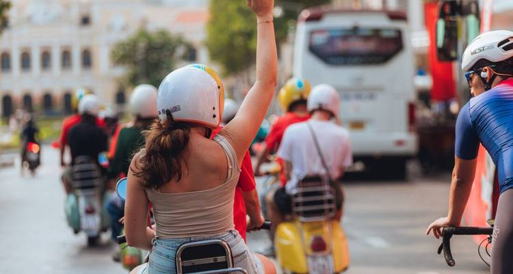 Scooterrijders met helmen scheren voorbij op stadsstraten, één reiziger steekt enthousiast een hand omhoog.